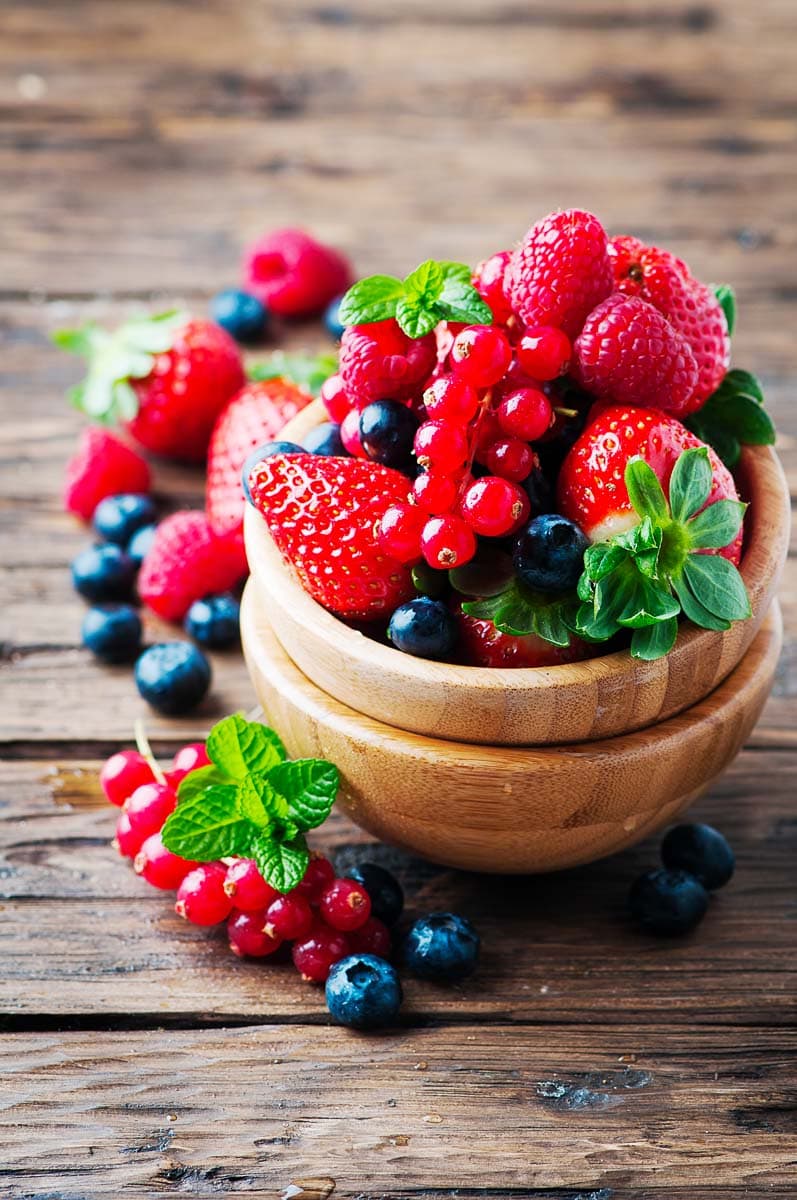 A wooden bowl filled with mixed berries including strawberries, blueberries, and red currants, garnished with fresh mint leaves on a rustic wooden surface.