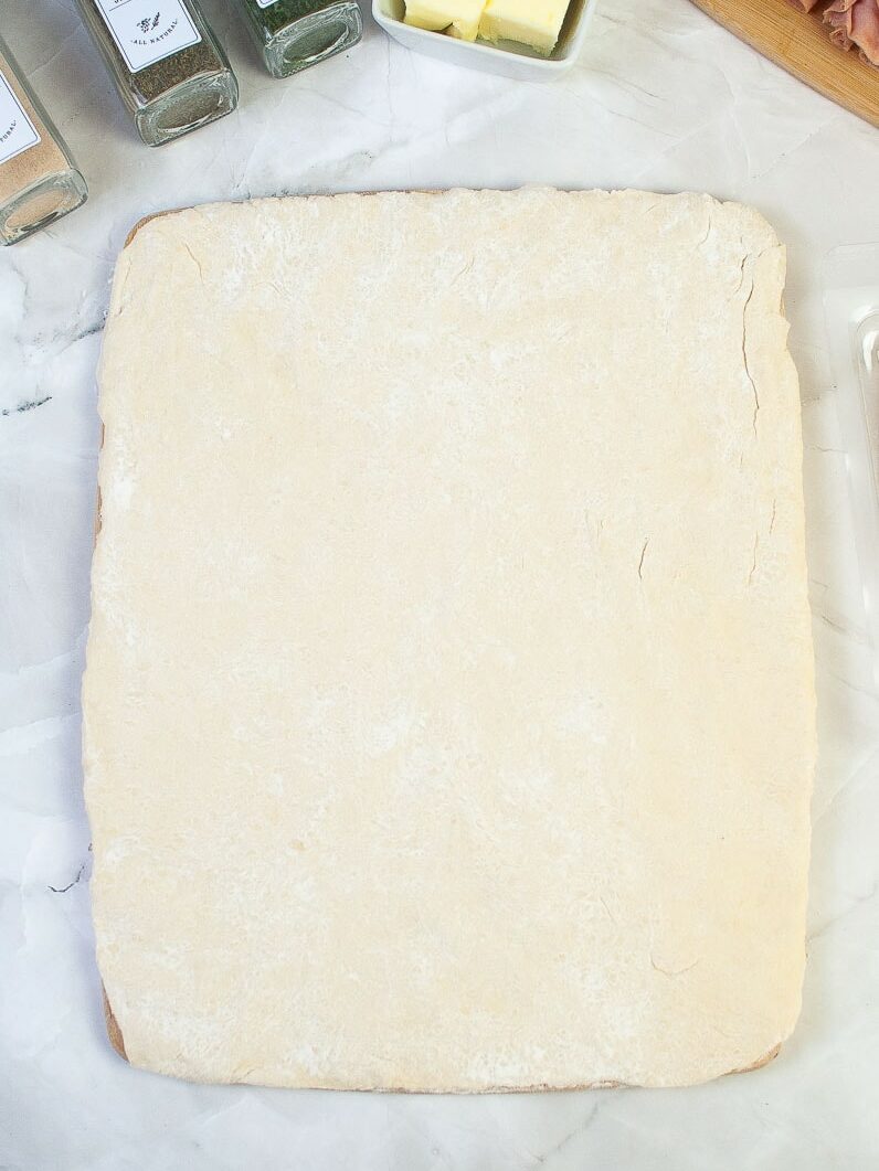 A sheet of raw pastry dough on a marble surface with ingredients like butter and ham in the background.