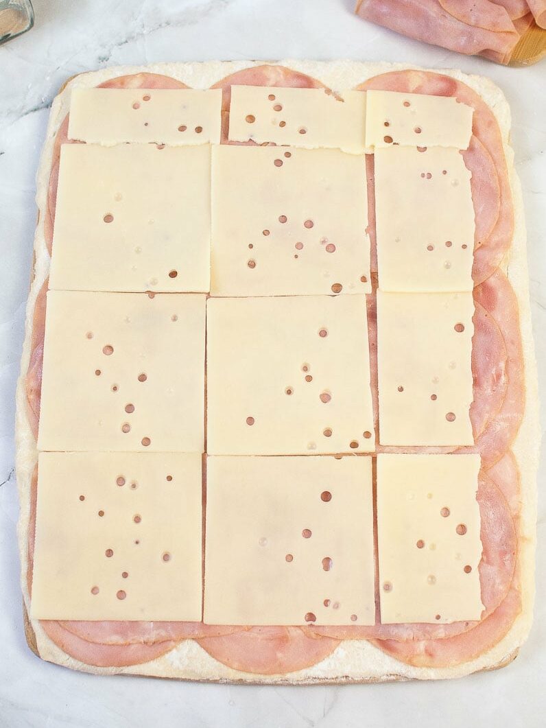 Unbaked pizza dough with ham slices topped with evenly arranged cheese slices.