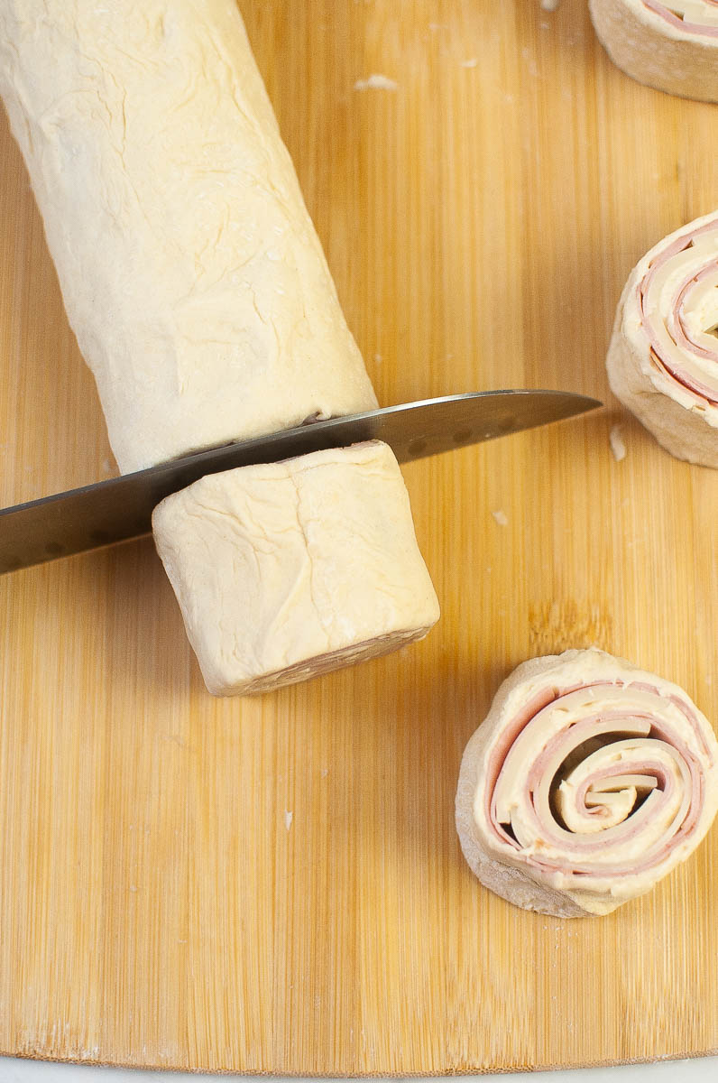 Slicing rolled dough to make ham and cheese pizza rolls on a wooden cutting board.