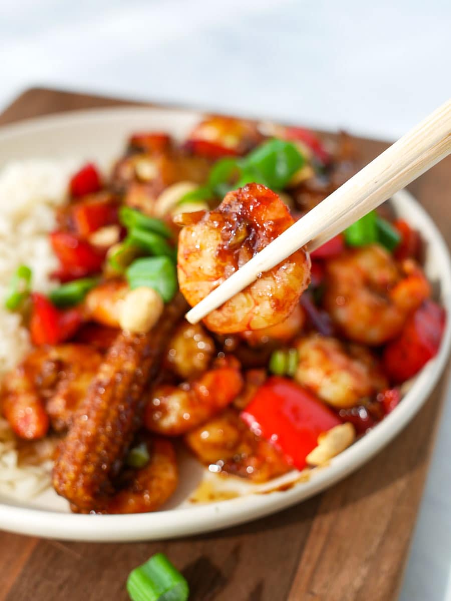 A bowl of shrimp stir-fry with vegetables served over rice, with one shrimp held by chopsticks.