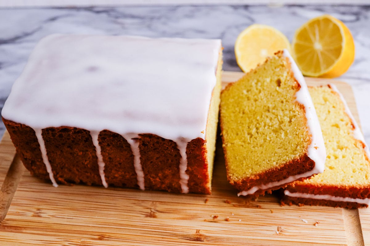 A lemon loaf cake with glaze on a wooden board with sliced lemons in the background.