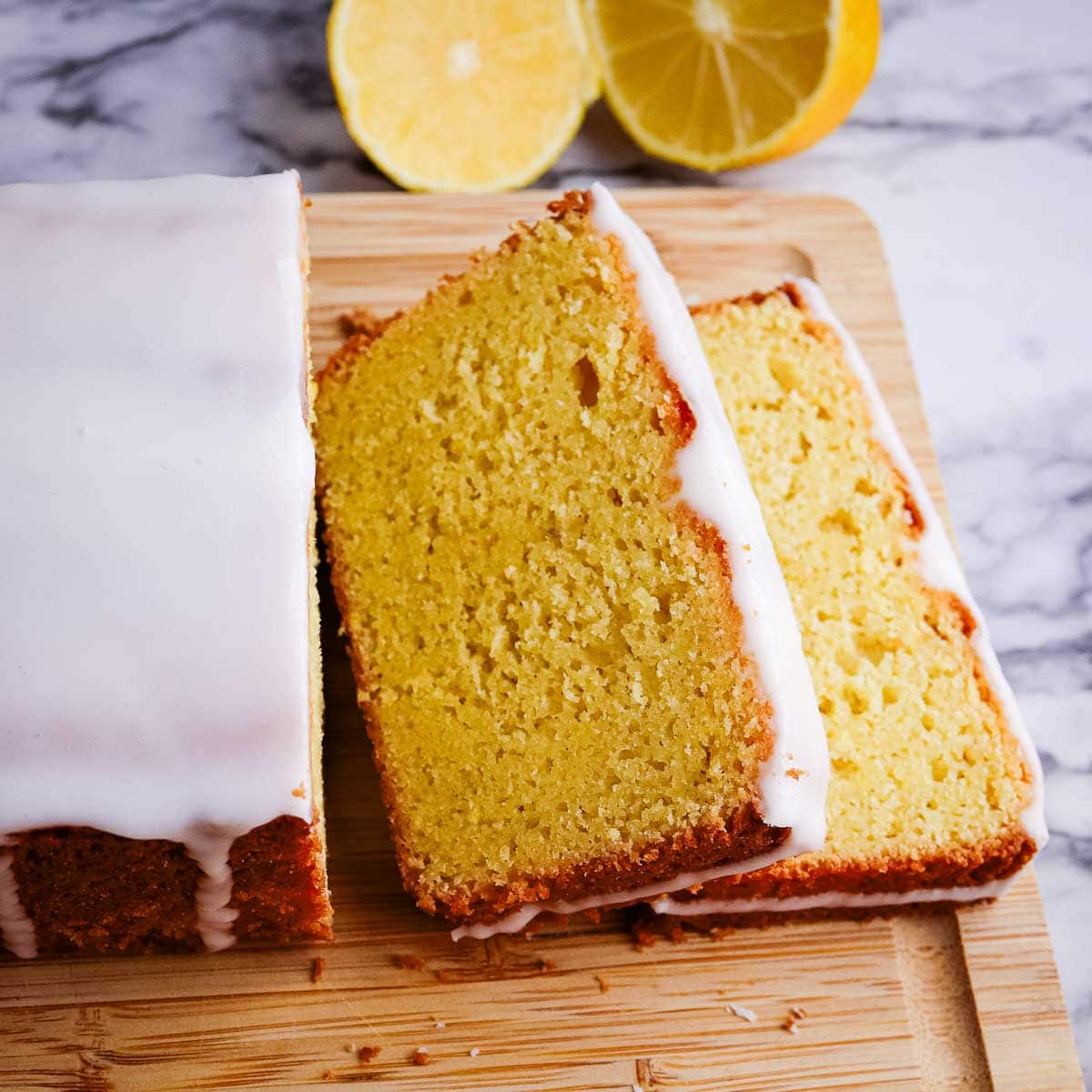 A lemon loaf cake with glaze on a wooden board with sliced lemons in the background.