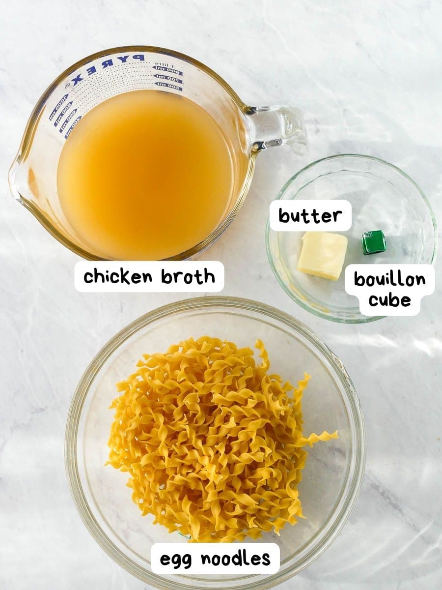 Ingredients for cooking: chicken broth, egg noodles, butter, and a bouillon cube on a kitchen counter.