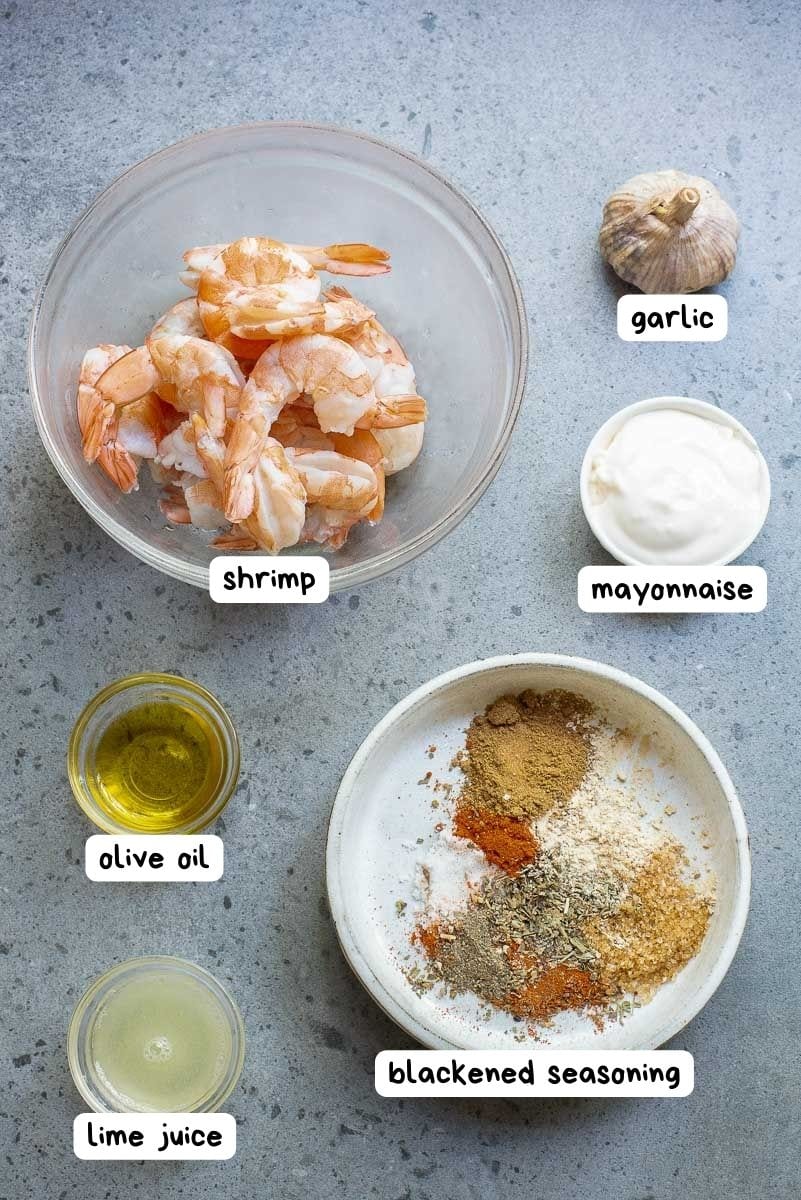 Ingredients for a recipe arranged on a counter: shrimp in a bowl, garlic, mayonnaise, olive oil, lime juice, and a bowl of blackened seasoning.
