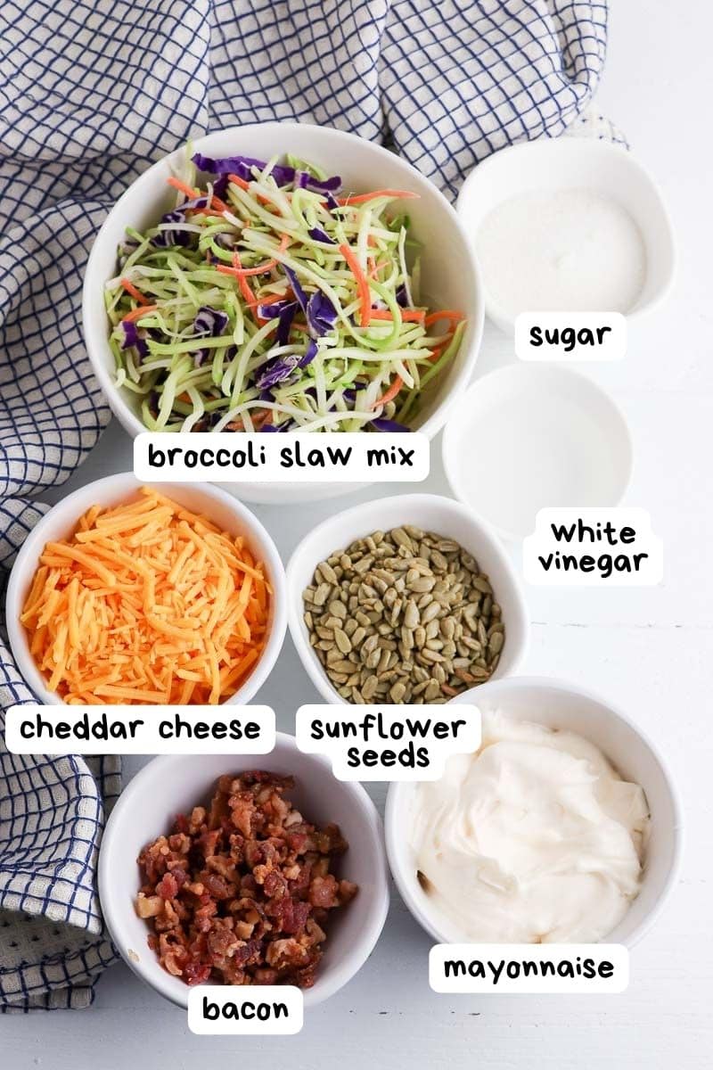 Bowls of ingredients for broccoli slaw including slaw mix, shredded cheese, sunflower seeds, bacon, mayonnaise, sugar, and vinegar arranged on a white cloth.