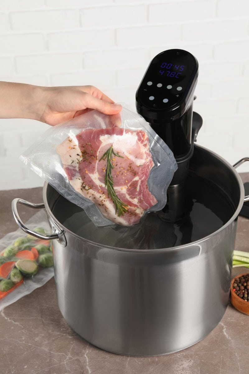 Person placing seasoned raw chicken thighs into a sous vide cooker with digital timer set for sous vide cooking.