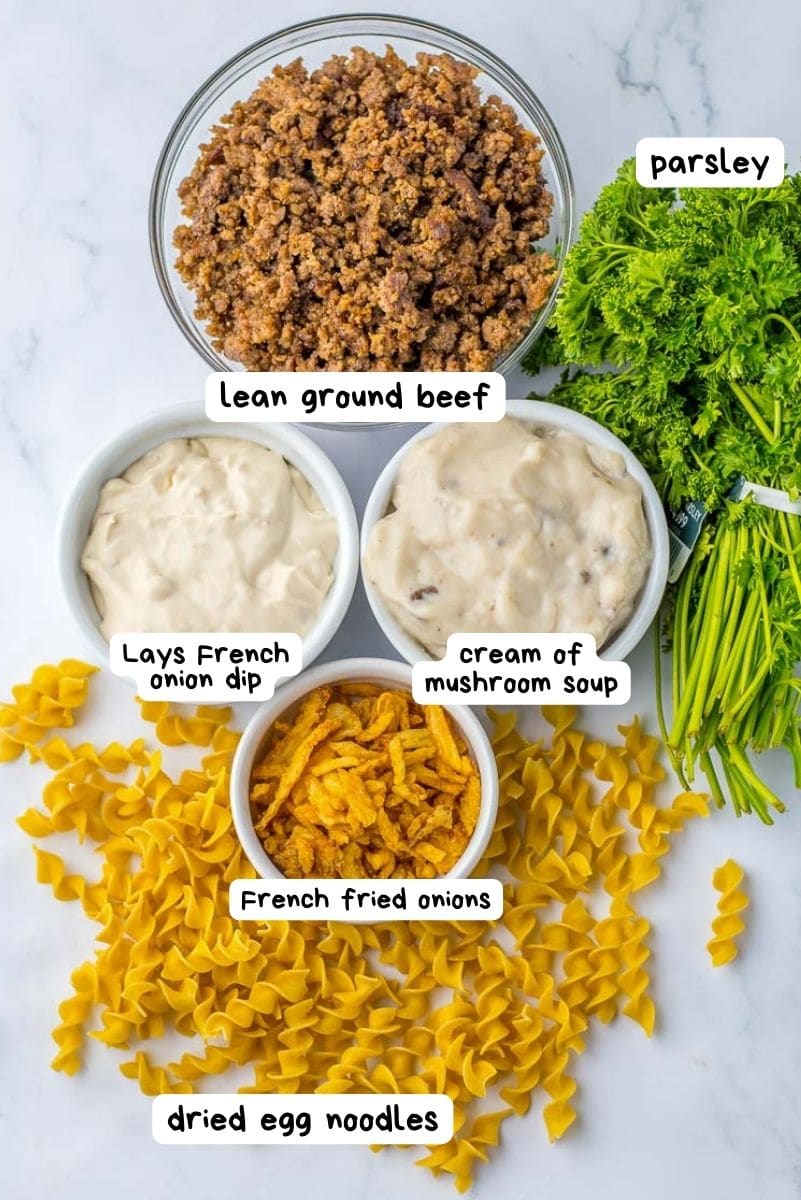 Ingredients for a French onion casserole dish laid out on a surface, including lean ground beef, french onion dip, cream of mushroom soup, french fried onions, dried egg noodles, and parsley.