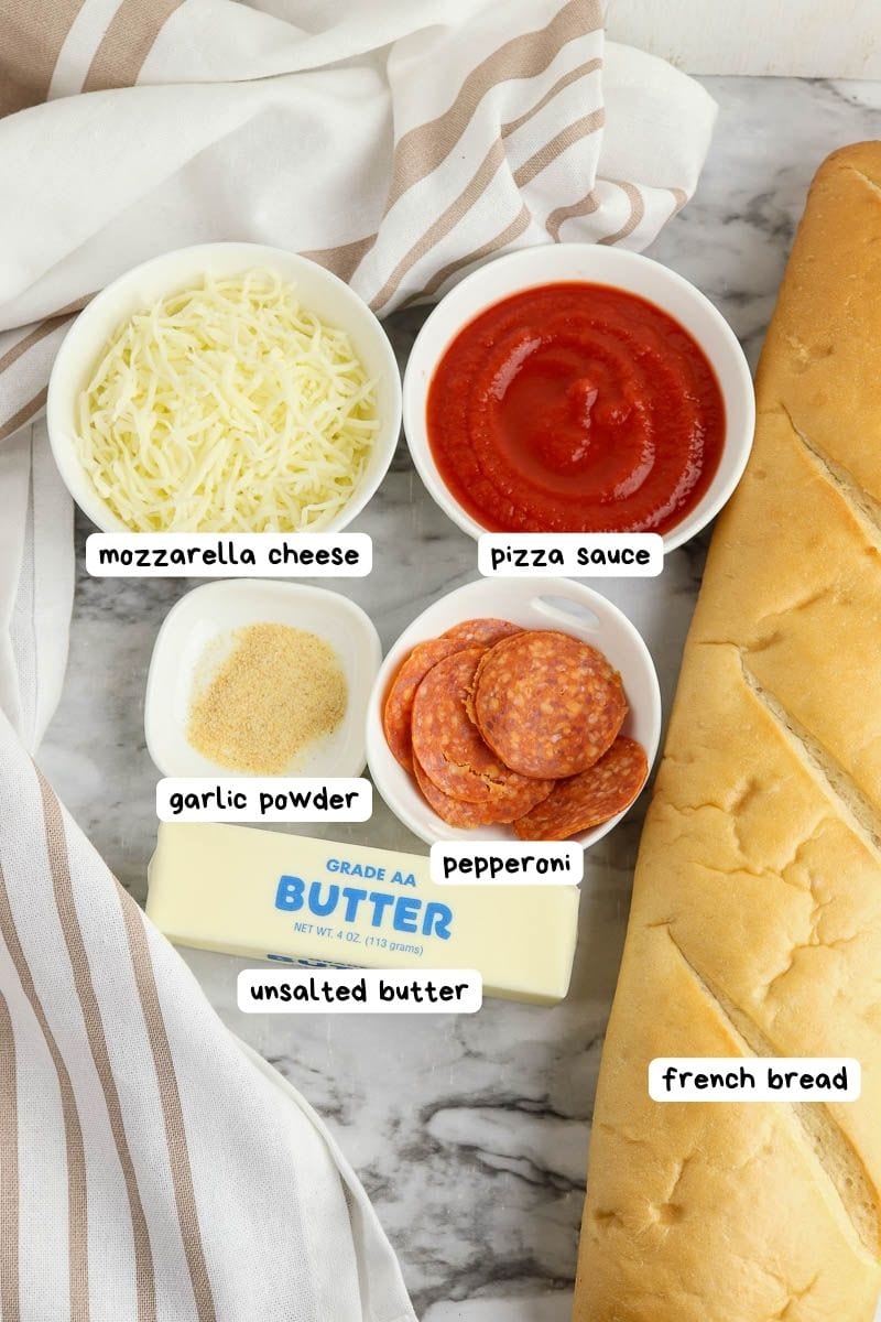 Ingredients for making pizza on french bread, including mozzarella cheese, pizza sauce, garlic powder, pepperoni, unsalted butter, and a loaf of french bread, all labeled on a kitchen counter.