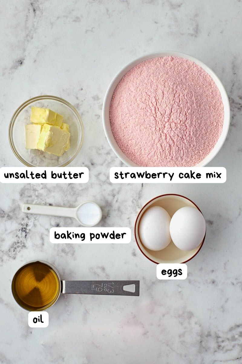 Ingredients for strawberry donuts preparation laid out on a marble surface, including unsalted butter, strawberry cake mix, baking powder, eggs, and oil, each labeled accordingly.
