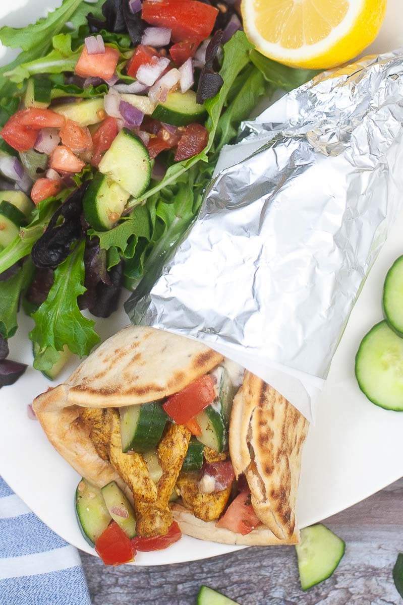 A chicken in a pita with fresh vegetables, wrapped in foil, served on a plate alongside a salad and cucumber slices.