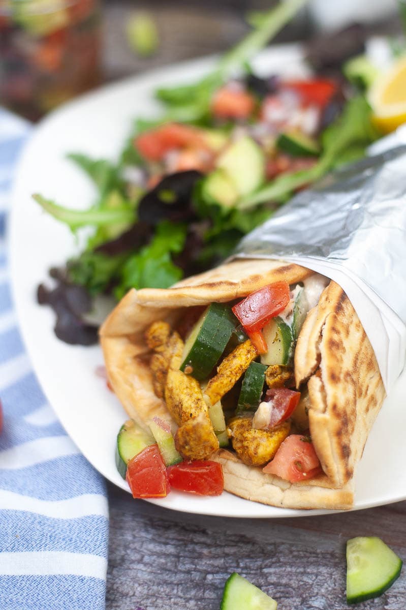 A chicken pita wrap filled with vegetables on a white plate, with a side salad and a blue and white napkin.