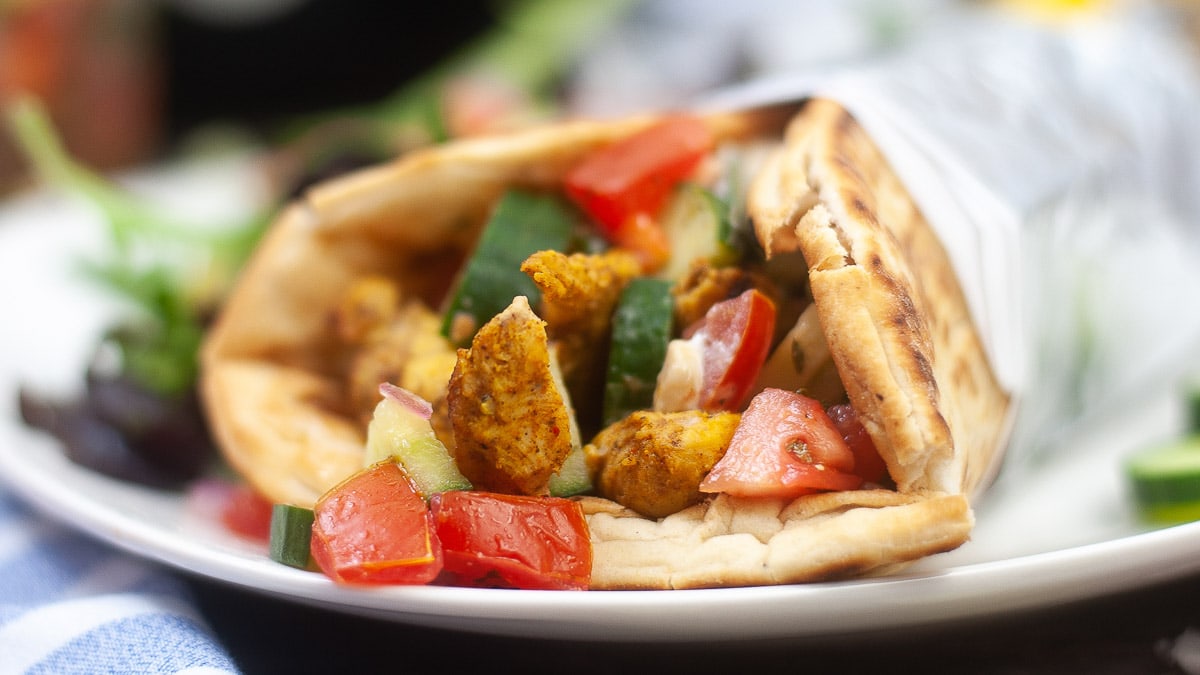 A close-up of a pita bread sandwich filled with spiced chicken, tomatoes, and green peppers, served on a white plate with a side salad.