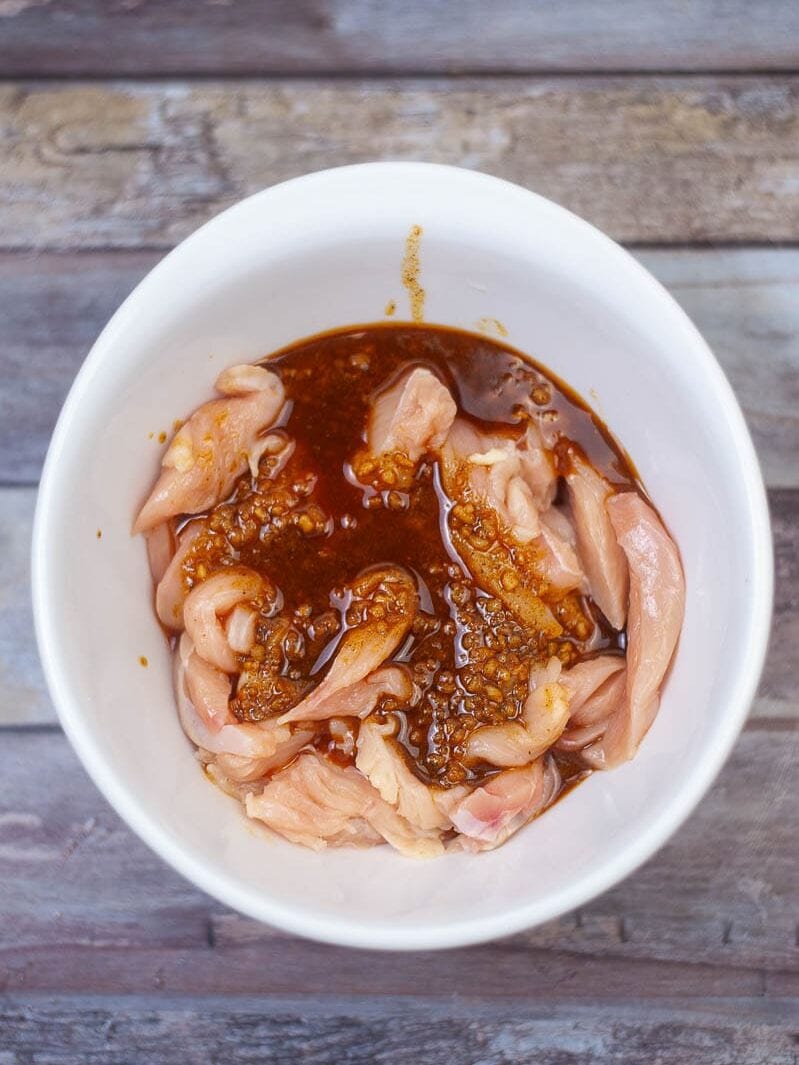 Raw chicken pieces marinating in a spicy red sauce in a white bowl on a wooden surface.