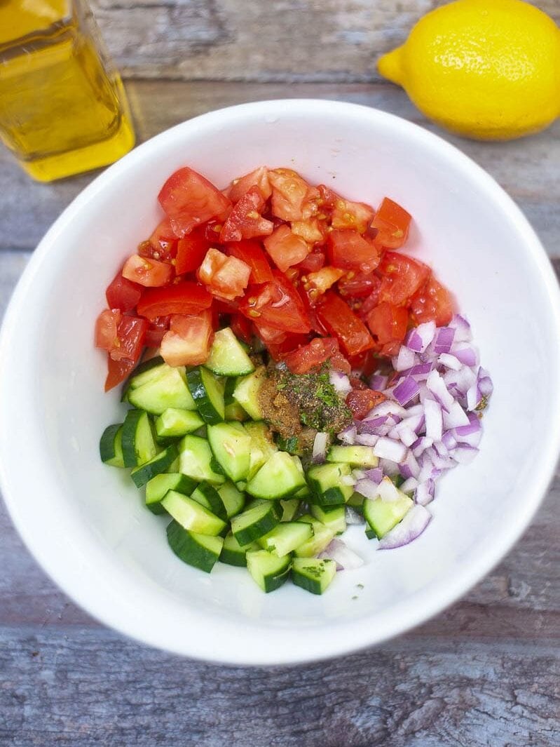 A bowl of chopped tomatoes, cucumbers, and red onions on a wooden table, with a lemon and oil bottle on the side.