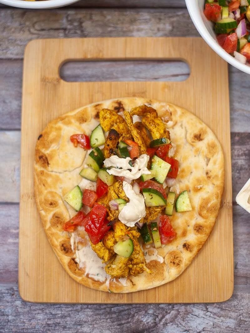 A flatbread topped with roasted chicken-spiced vegetables, diced tomatoes, cucumbers, and a dollop of white sauce, served on a wooden cutting board.
