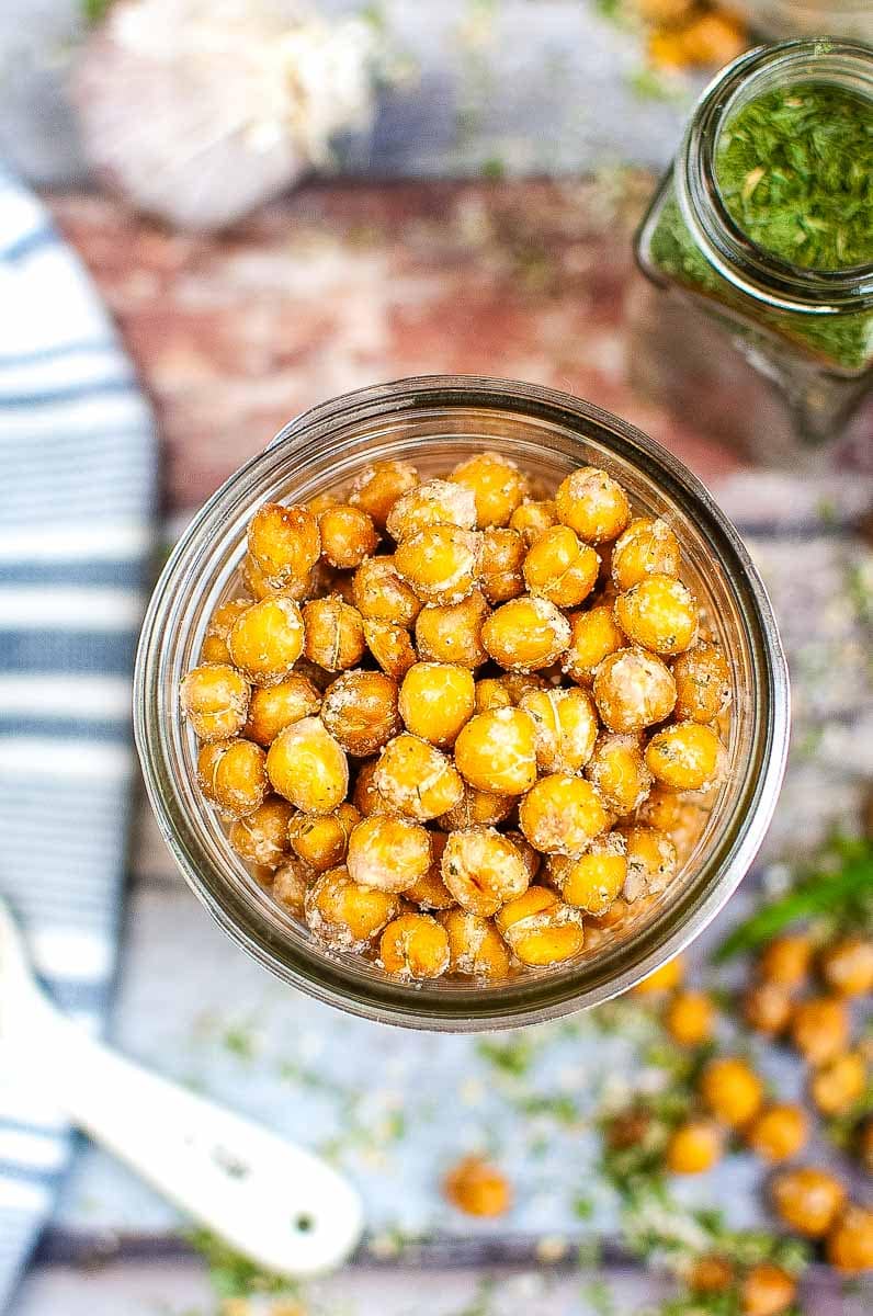 Top view of a jar of roasted chickpeas sprinkled with seasoning, with a spoon and more chickpeas visible in the background.