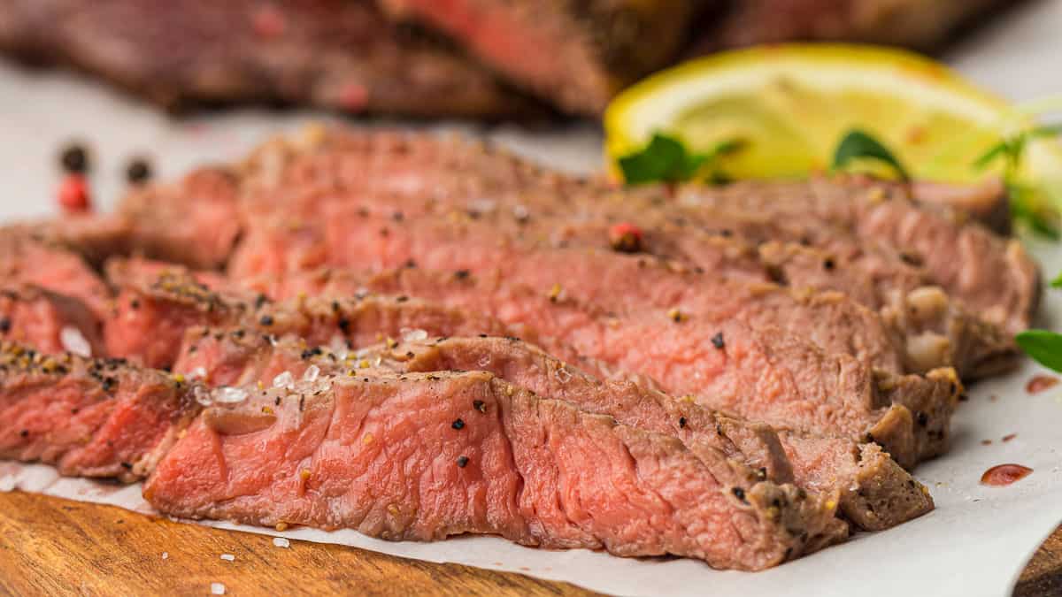 Slices of medium-rare steak seasoned with pepper and salt on a wooden board, garnished with herbs and a lemon wedge in the background.