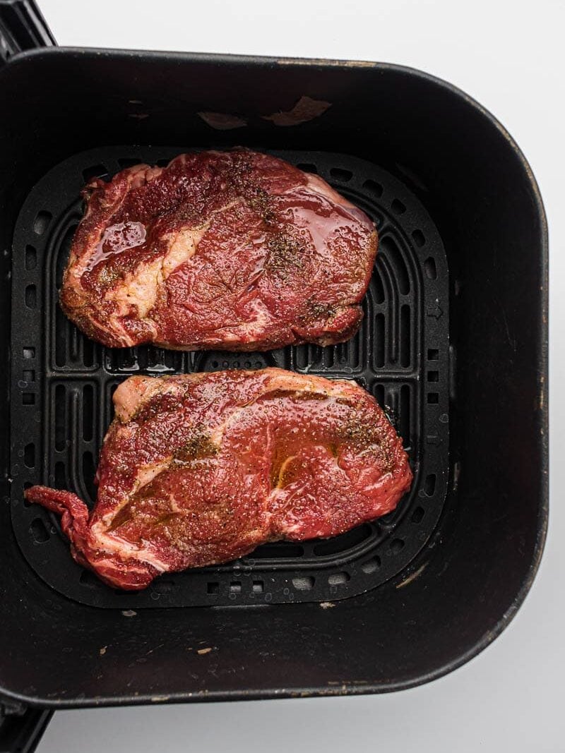 Two seasoned ribeye steaks in an air fryer basket ready to be cooked to perfection.