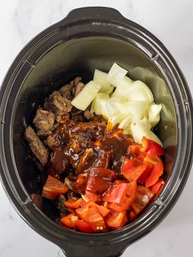 Ingredients for a meal inside a slow cooker, including beef, tomatoes, and onions, prior to cooking.