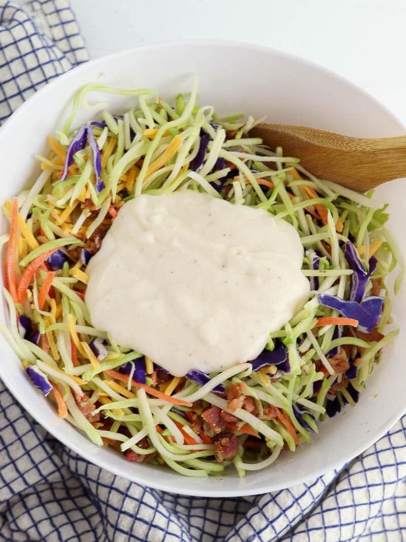 A bowl of broccoli slaw with carrots, purple cabbage, and bacon, topped with a creamy dressing, on a blue and white checkered cloth.