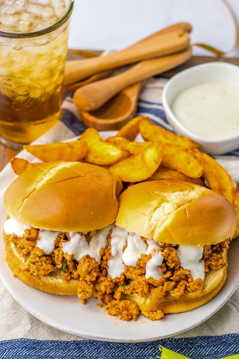 Two Buffalo Chicken Sloppy Joe sandwiches with creamy sauce on a plate with potato chips and a glass of iced tea in the background.