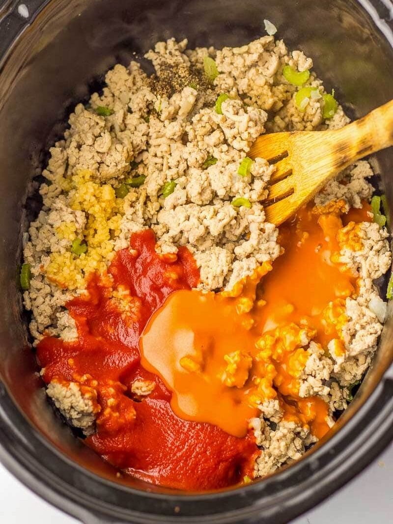 Ground chicken, diced vegetables, and tomato sauce in a slow cooker, stirred by a wooden spoon.