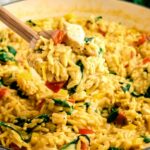 A wooden spoon scoops up a serving of creamy chicken and vegetable orzo from a large pot.