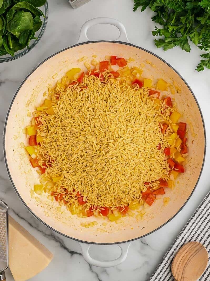 A pot containing uncooked orzo pasta mixed with chopped red and yellow bell peppers on a kitchen counter, surrounded by ingredients like spinach, parsley, salt, and pepper.