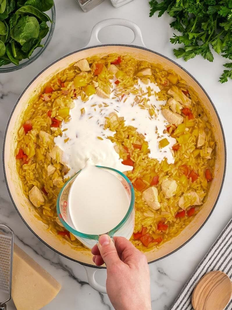 A hand pouring half and half into a skillet with chicken, rice, bell peppers, and herbs on a kitchen counter surrounded by fresh ingredients.