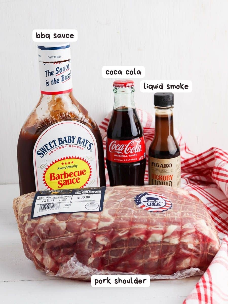 A pork shoulder wrapped in plastic on a table, accompanied by a bottle of bbq sauce, coca-cola, and liquid smoke, all labeled for a cooking recipe.