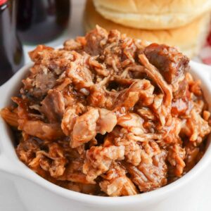 A bowl of pulled pork with barbecue sauce, served with hamburger buns in the background.