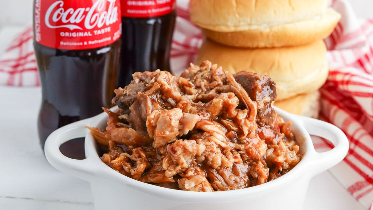 A serving of pulled pork in a white bowl with hamburger buns and bottled coca-cola in the background on a checkered cloth.