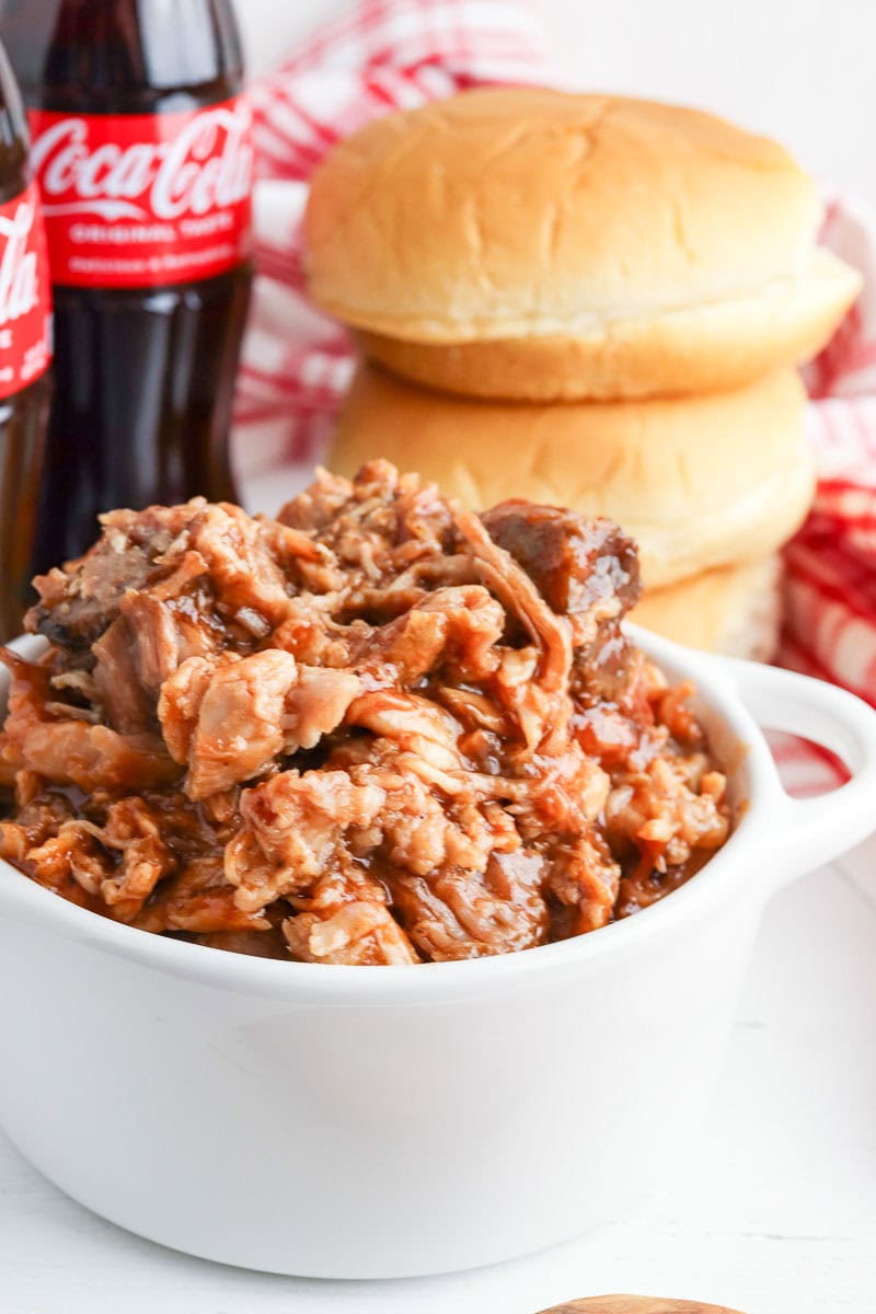 A bowl of pulled pork served next to hamburger buns and coca-cola bottles, with a red and white checkered napkin in the background.