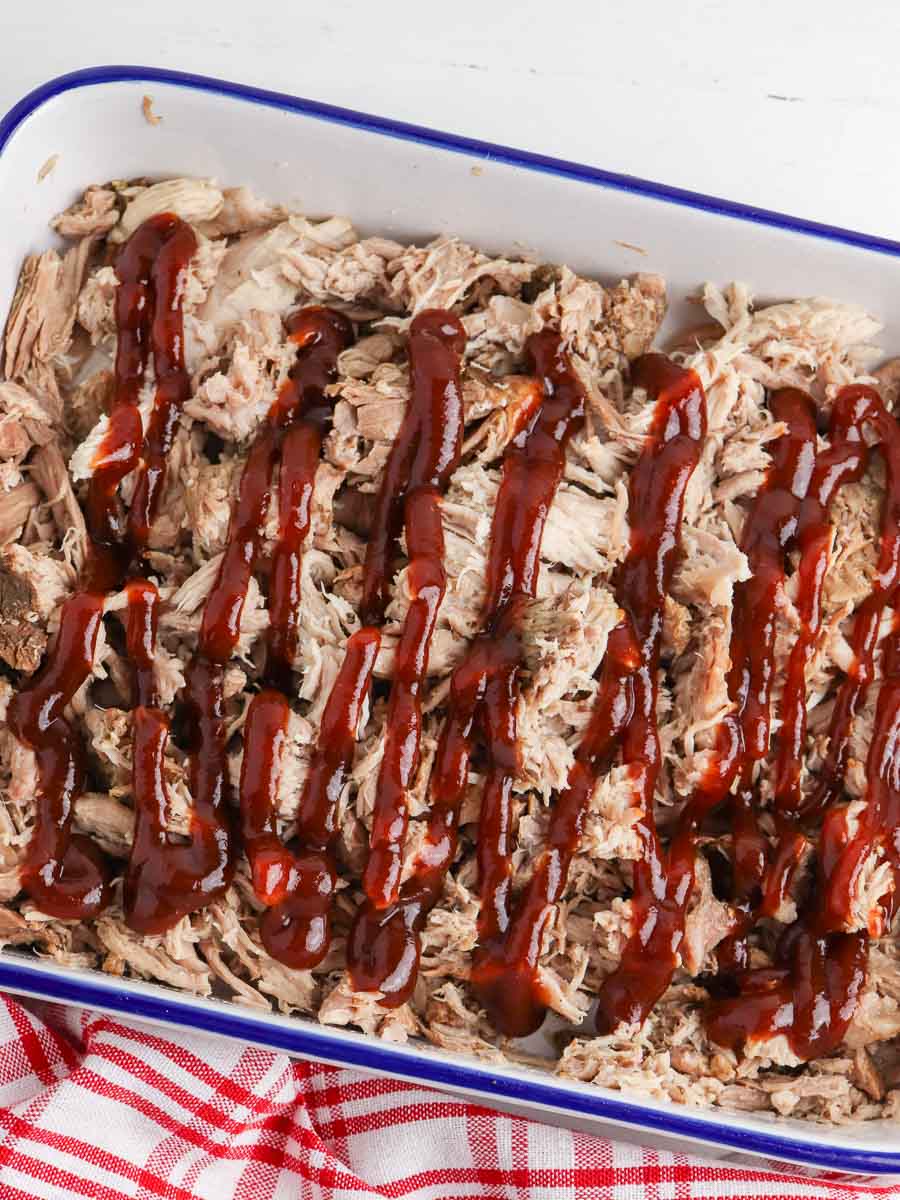 Shredded roast pork in a blue baking dish, drizzled with barbecue sauce, on a red and white checkered cloth.
