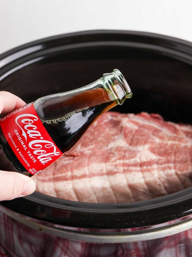 A hand pouring coca-cola from a bottle onto a slab of raw pork in a slow cooker.