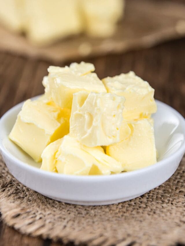 Chunks of fresh butter in a small white bowl on a wooden table with a burlap cloth underneath.