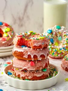 Stack of three pink frosted donuts with colorful sprinkles on a white plate, with a glass of milk in the background.