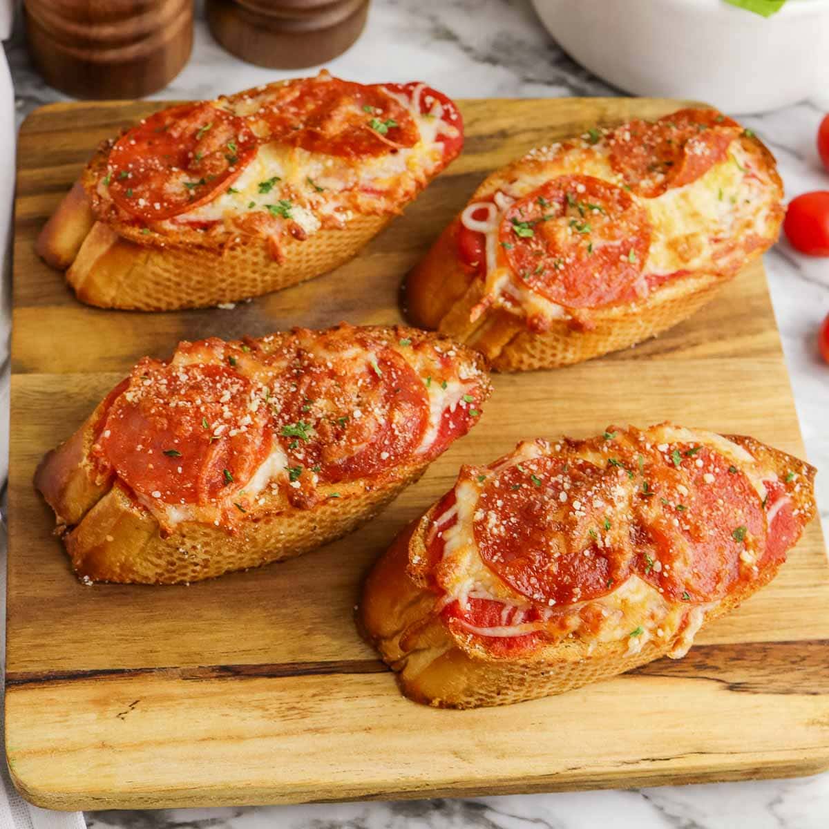 Four pepperoni pizza toasts on a wooden cutting board, garnished with herbs and served on a marble countertop.