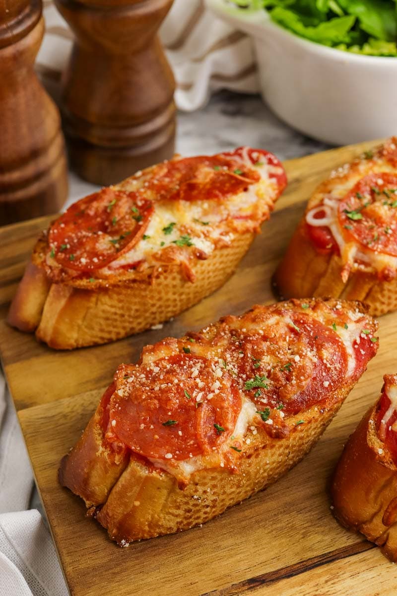 Three slices of pepperoni pizza baguette on a wooden cutting board, garnished with parsley, beside salt and pepper shakers and a bowl of salad.