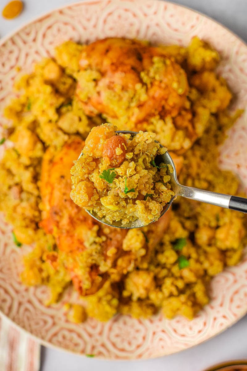 A bowl of quinoa and Moroccan chicken thighs with tomatoes and peas, with a spoon scooping some out, showcasing a close-up of the dish.