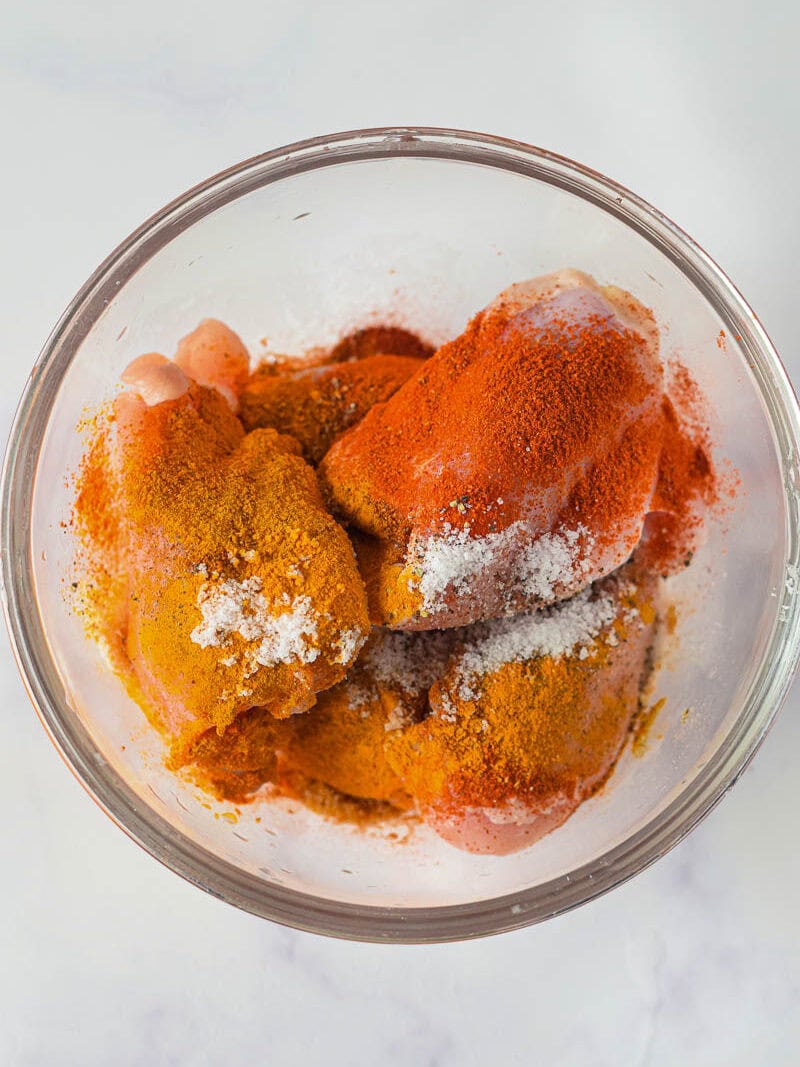 Raw Moroccan chicken thighs in a clear bowl with orange and red spices ready for marinade.