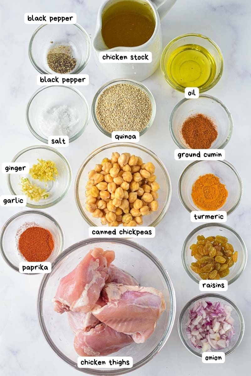 Ingredients for a recipe displayed on a white surface, labeled individually, including spices, oil, quinoa, chickpeas, Moroccan chicken thighs, and chopped onions.