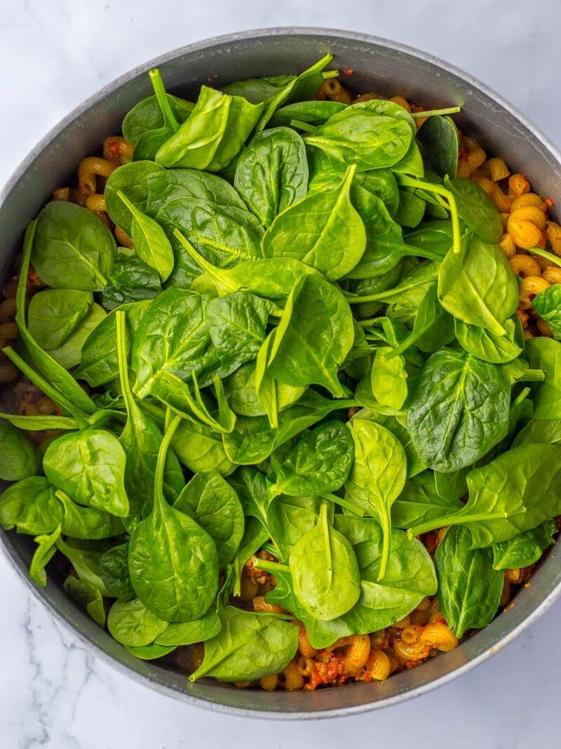 Fresh spinach leaves on top of cooked pasta in a gray pot, set on a white marble countertop.