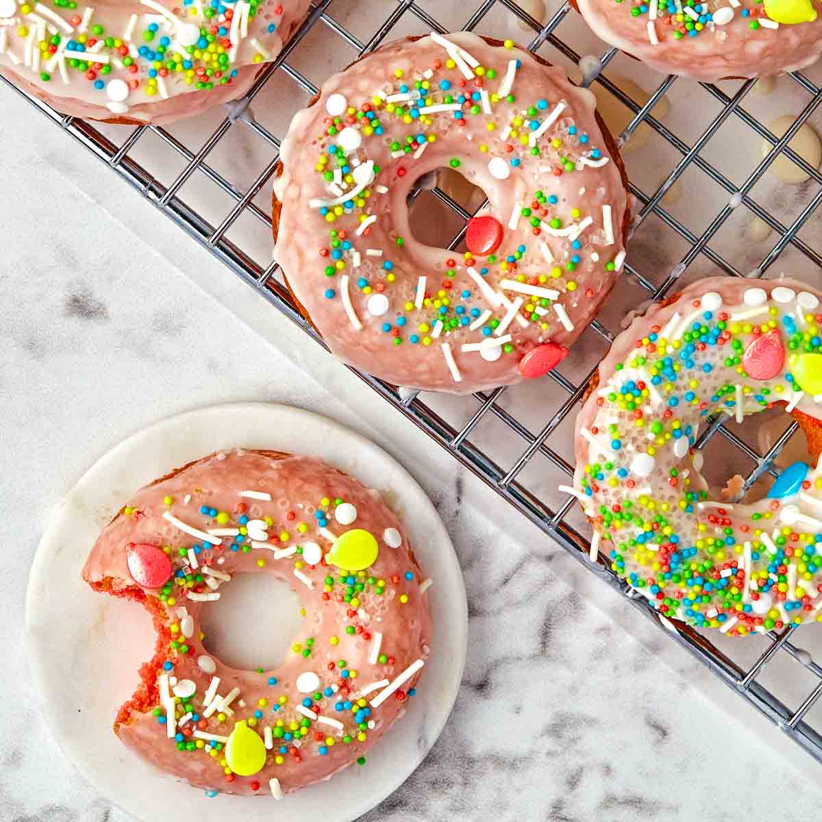 Colorful sprinkle-topped donuts with pink icing on a cooling rack and plate.