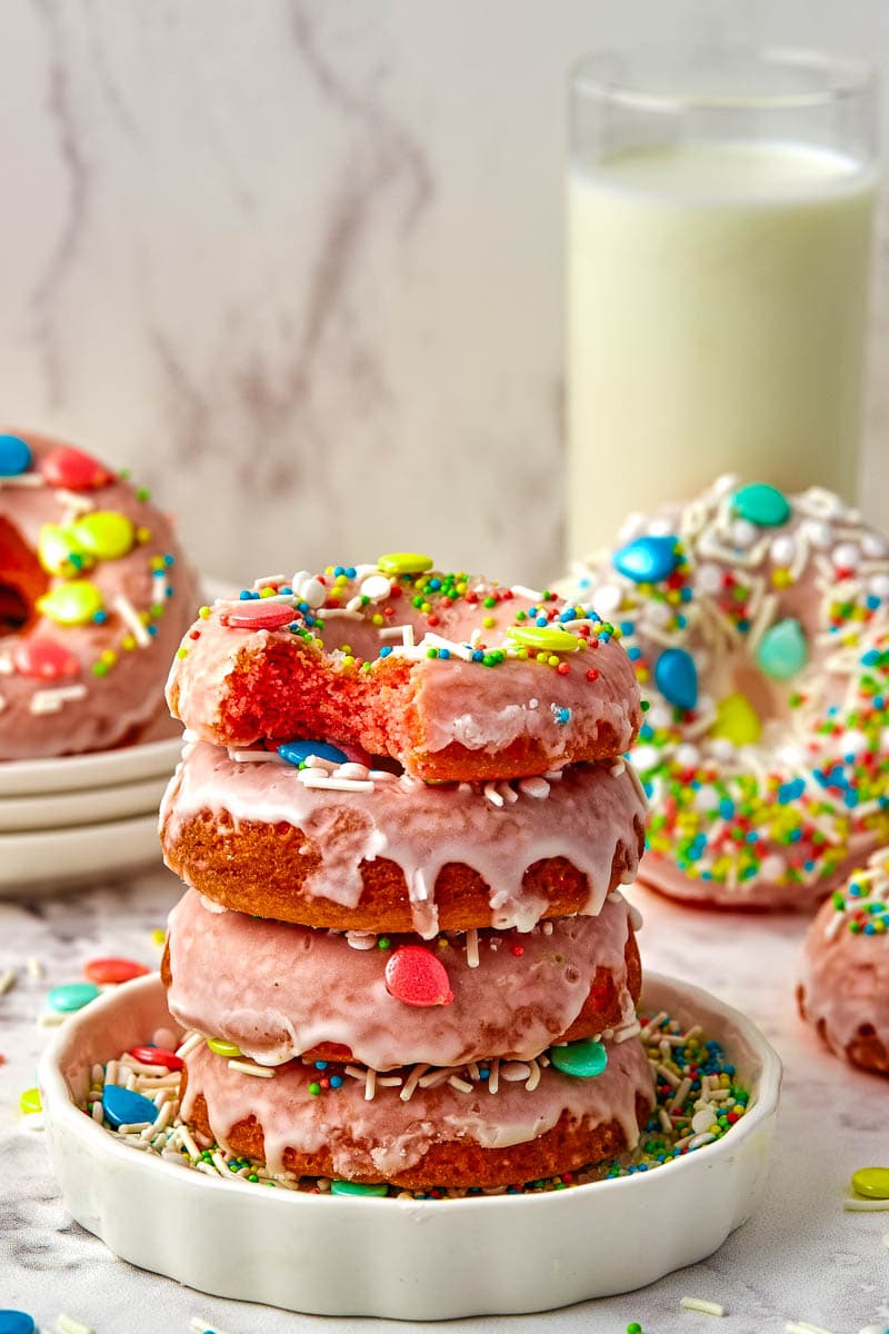 A stack of pink frosted doughnuts with colorful sprinkles, accompanied by a glass of milk in the background.