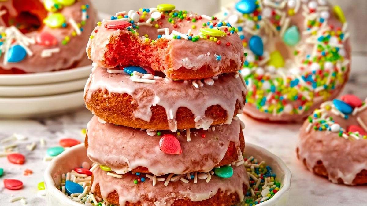 A stack of colorful, glazed doughnuts with sprinkles, displayed on a white surface surrounded by scattered candies.