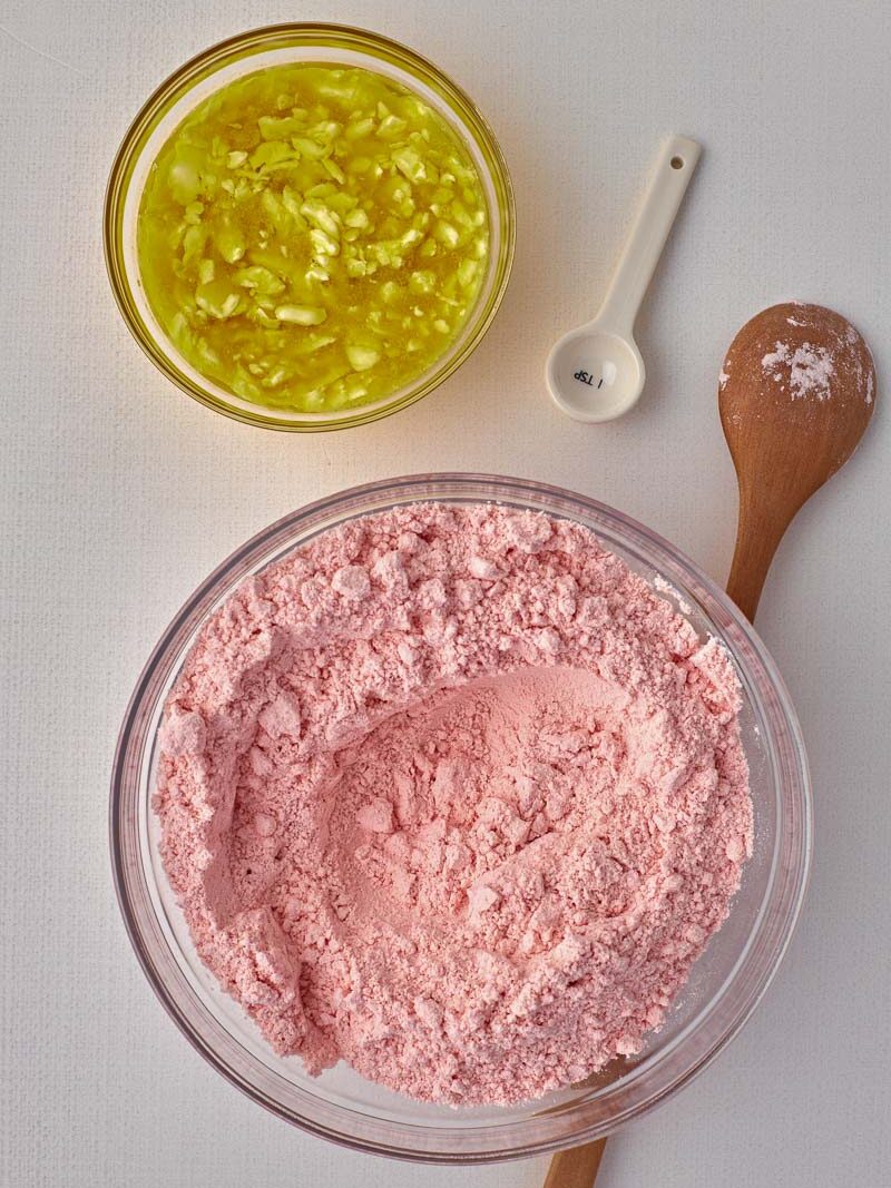 Ingredients for baking on a table: a jar of chopped nuts, a bowl of pink flour mixture, a measuring spoon, and a wooden spoon sprinkled with flour.