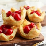 Cherry pastries arranged on a wooden board, featuring flaky golden crusts filled with vibrant red cherries.