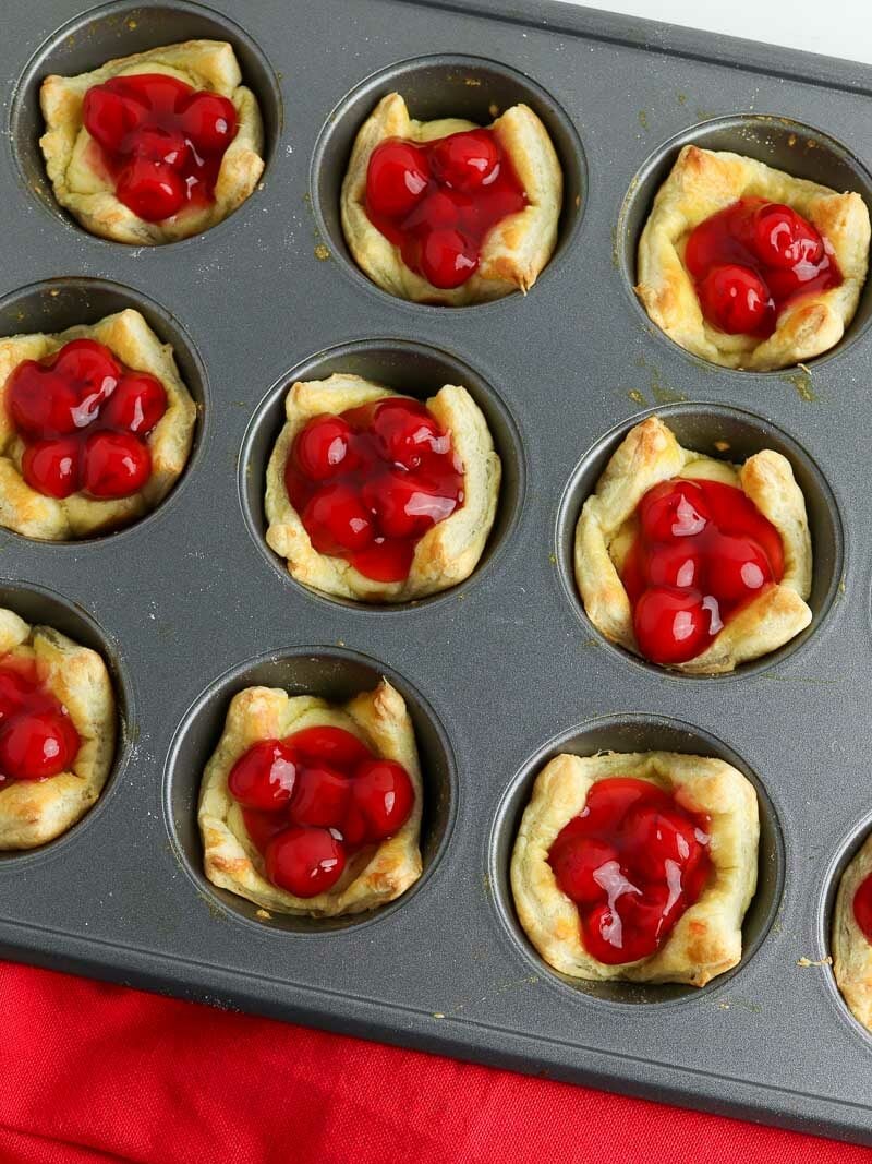 Freshly baked cherry pies in a muffin tin viewed from above, with a red napkin partially visible.
